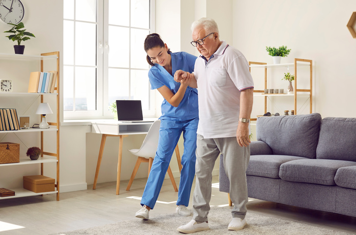 Young nurse helping elderly man walk in the room, holding his hand, supporting him.