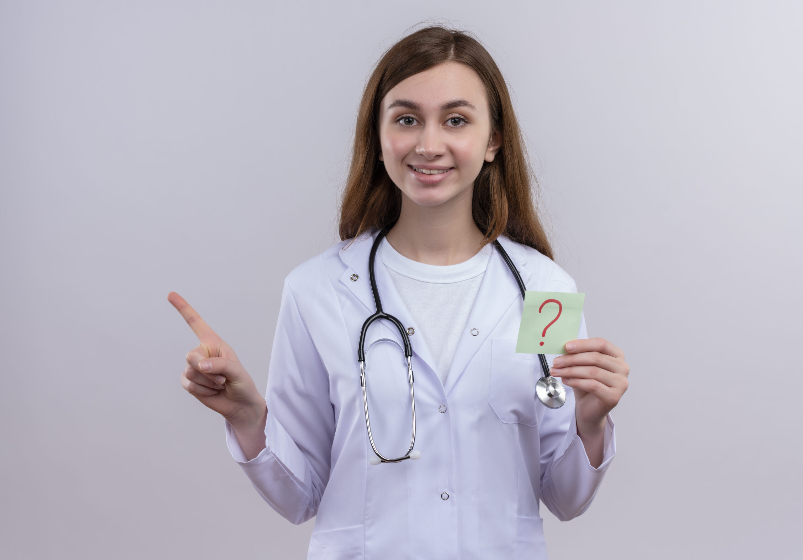 smiling young female doctor wearing medical robe and stethoscope holding paper note with question mark written on it and pointing at left side on isolated white background